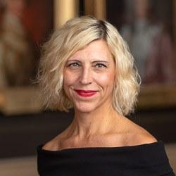 headshot of Ulrika Dahl - a white woman with a jaw-length, razored bob of wavy, silvery blonde hair, and a playful expression. She is wearing berry red lipstick and a black boat-neck top.